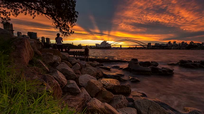 A rocky shoreline with tourists visiting the coast with a city skyline in the distance as sunset turns into night