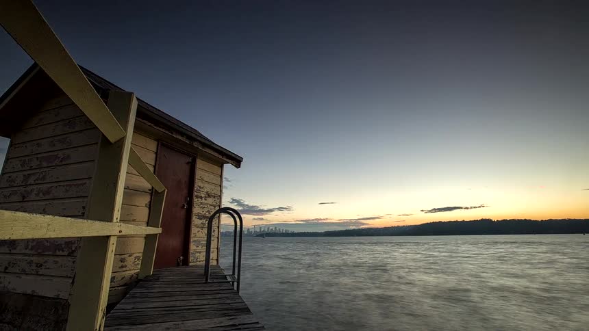 A time lapse view shows a small weathered wooden hut on a pier beside a lake as orange and white clouds move across the sky during a sunset that fades into a dark blue evening
