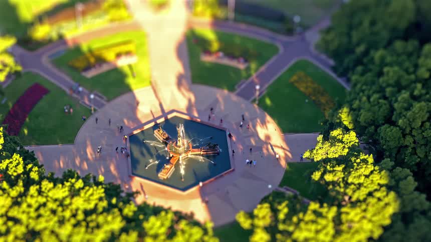An aerial tiltshift view shows people gathered around a central fountain in a park with intersecting pathways and surrounding trees