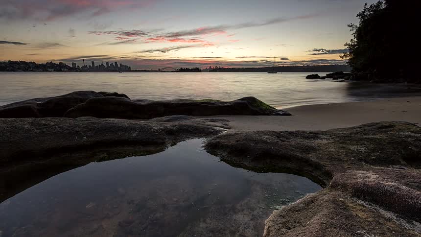 Rocks and calm seawater by the shore with an urban skyline at dusk