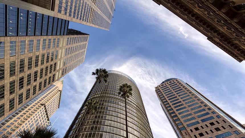 The video features several highrise buildings reaching toward a blue sky with scattered clouds filmed from a lowangle perspectivethin palm trees are interspersed between the modern glass and stone structures which shift as the camera pans upward an