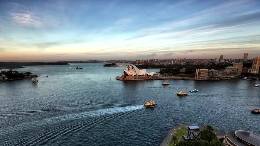 Boats sail off the coast near the sydney opera house as traffic moves in the city with buildings and skyscrapers