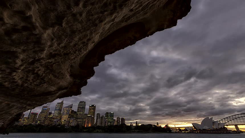 A glowing city skyline and the sydney opera house appear across the water beneath dramatic clouds and a rocky overhang at dusk
