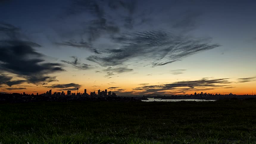 A wide view shows a city skyline and bridge at sunset beneath dramatic clouds seen from a grassy hill
