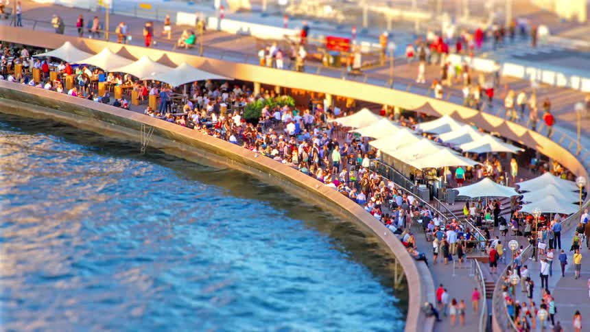 A tilt shift view shows a high angle of a crowded waterfront promenade with many people walking and sitting at outdoor cafes under white awnings next to blue harbor water on a bright sunny day
