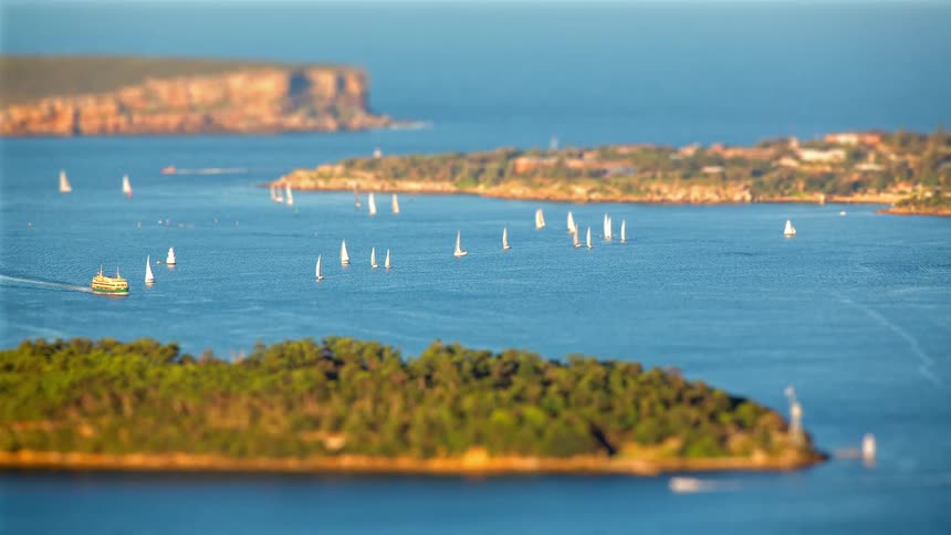 A tiltshift view shows sailboats scattered across a bright blue harbor with islands and coastal cliffs in the distance