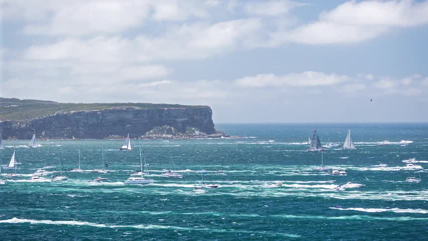 Many sailboats and yachts navigate choppy blue water near a massive coastal cliff under a cloudy sky while smaller motorboats and a red ferry cruise through the busy harbor during a daytime maritime event