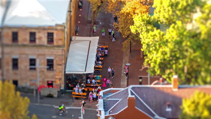 People sitting at tables and walking on the street in front of neighborhood buildings as trees move in the breeze