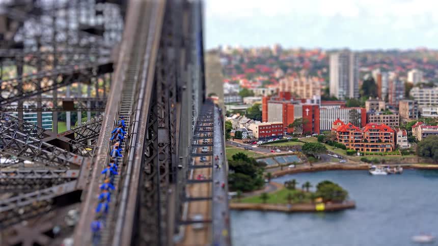 A tilt shift view of a steel bridge with traffic moving toward a waterfront city and harbor