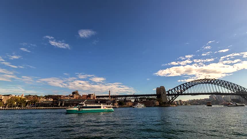 This wideangle panning shot showcases the sydney harbour skyline featuring bustling ferry traffic and the iconic harbour bridge the camera moves along a sundrenched waterfront walkway to reveal the white sails of the opera house under a b