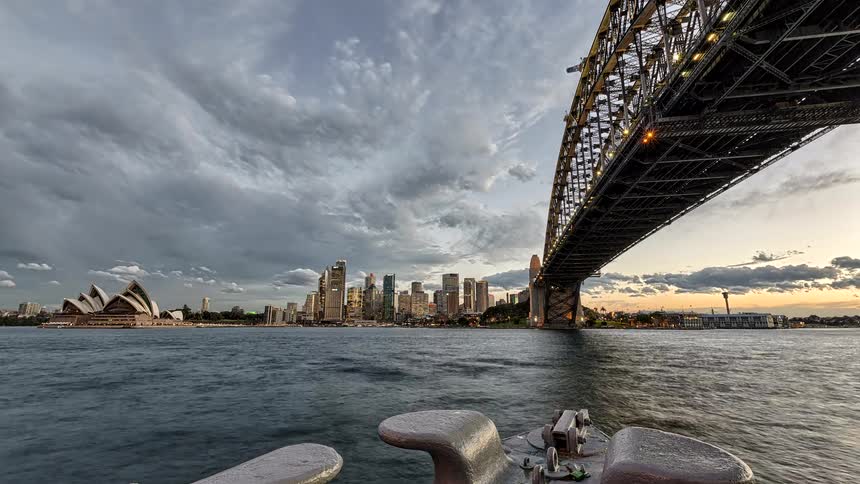 A time lapse view from a boat in sydney harbour shows the sun setting over the city skyline as lights begin to glow on the sydney opera house and the sydney harbour bridge under a moving cloudy sky