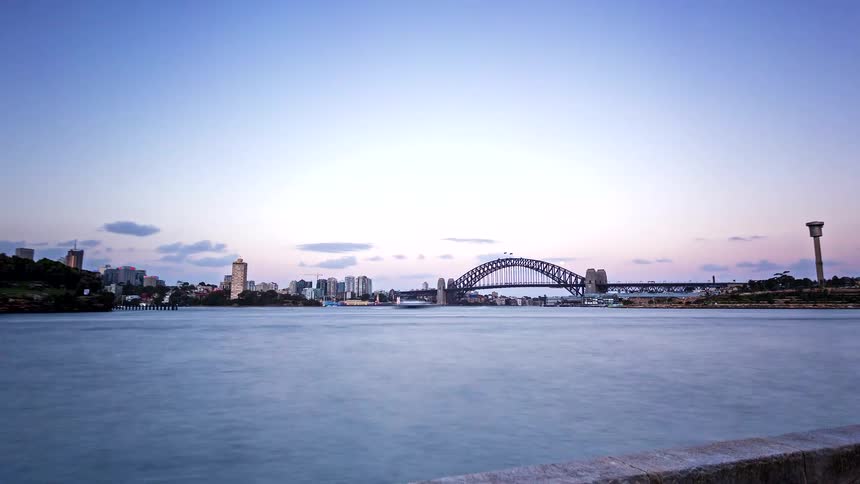 A time lapse view shows a large arched bridge over blue water as a purple sunset sky darkens into night and a bright moon rises behind the city skyline and harbor