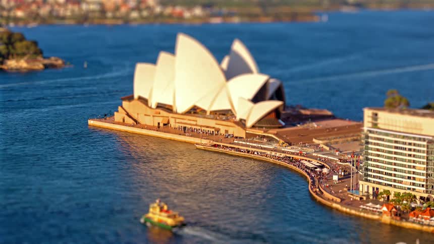 A large building stands on a paved peninsula next to deep blue water where a yellow and green ferry travels past a large crowd of people gathered along the shoreline walkway near a modern glass building