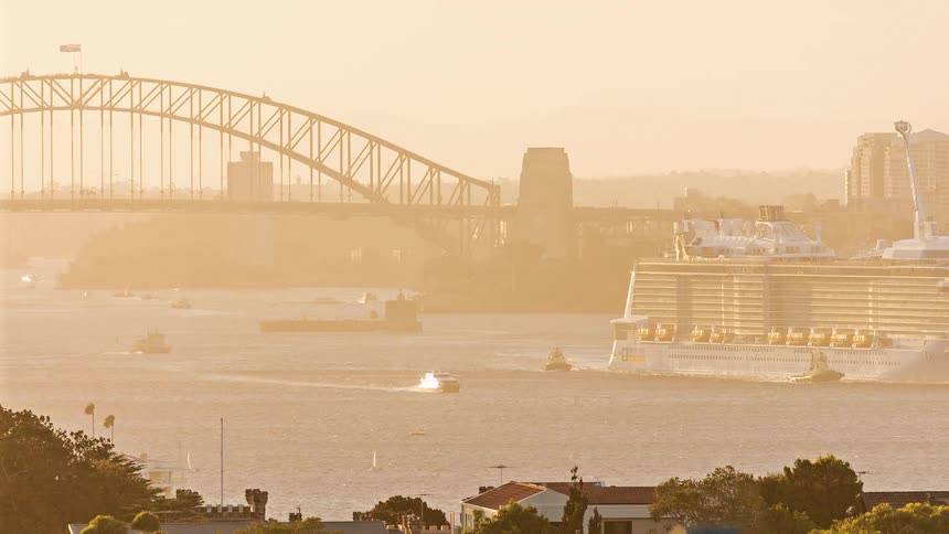 A large cruise ship sails through a harbor away from a steel arch bridge in warm evening light