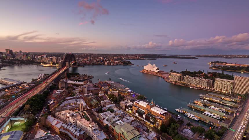 This aerial timelapse captures the transition from sunset to night over sydney harbour showing the city lights flickering to life the harbour bridge and opera house remain central as traffic trails and ferry movements create a vibrant glow