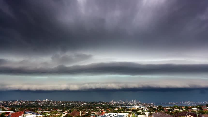 Dark heavy storm clouds rapidly roll over a coastal city skyline obscuring the sky with a dramatic shelf cloud formationrain begins to fall in visible sheets over the urban landscape as the dense cloud layer sweeps across the horizon