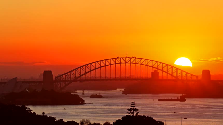 The sun shines brightly in the sky as boats sail under a bridge as traffic moves and the sun sets on the horizon