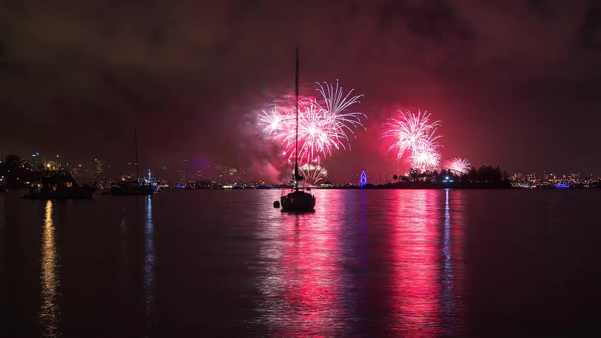 Boats sail on the water and metropolitan city lights glow as colorful fireworks explode across the night sky