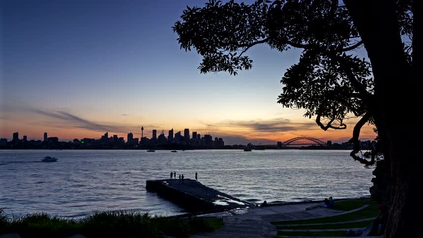A city skyline on the other side of a river with a bridge stretching over the water toward the city and the orange setting sun in the distance