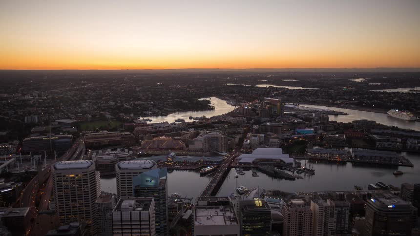 An aerial view shows a city and harbor at sunset with the sun setting on the horizon and rivers winding through the urban landscape