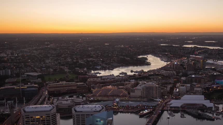 A time lapse view shows a high angle of a harbor city as the bright sun sets over the horizon and thousands of city lights flicker on across the buildings and streets during a clear transition from day to night