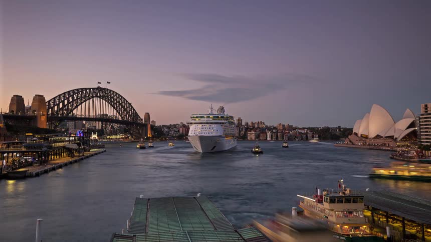 A large cruise ship sails through a busy harbor near a city skyline and iconic waterfront building during sunset