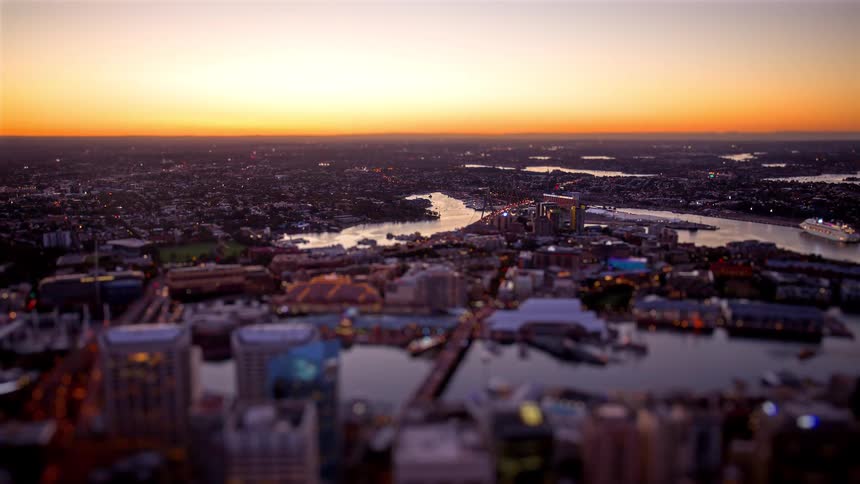 An aerial view of a sprawling city near a river that stretches toward the horizon at sunrise where the glowing orange sun rises above winding rivers bridges and densely packed buildings illuminated by the soft early morning light