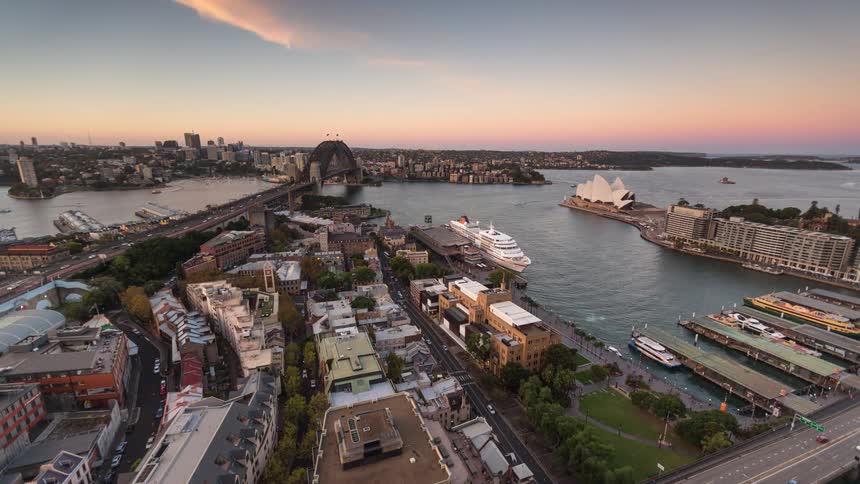 An aerial view of the iconic sydney harbour skyline featuring the sydney opera house and sydney harbour bridge with ships docking and sailing and cars moving across the busy bridge as day turns to night