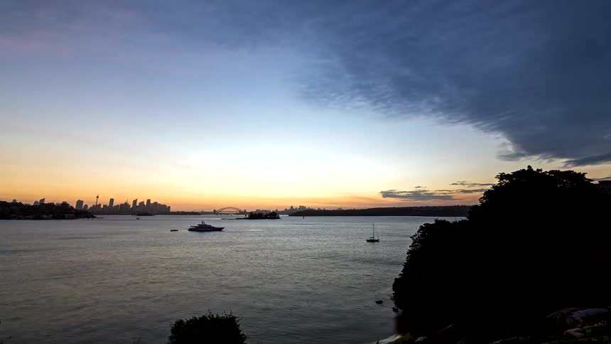 A time lapse view shows a bright sunset over a calm harbor with silhouettes of boats and a distant city skyline as pink clouds move across the sky and fade into a dark blue night