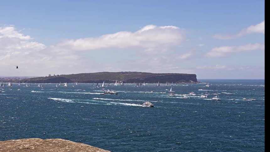 Many sailboats on the water off the coast as airplanes fly across a cloudy sky