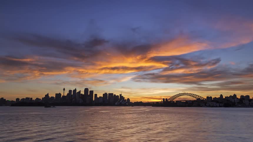 Boats sail on the water off the metropolitan coast with towers and skyscrapers on the city skyline and a bridge into downtown illuminated with lights as darkness falls