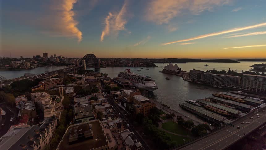 An aerial view shows a harbor with a cruise ship a large bridge and a waterfront city skyline in warm evening light