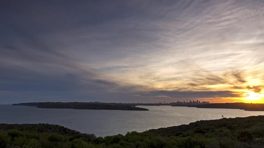 A wide view shows a harbor boats and distant city skyline at dusk under a dramatic cloudfilled sky