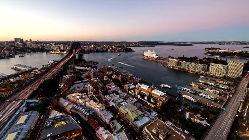 High angle time lapse shows the sun setting over sydney harbour as city lights and traffic on the harbour bridge illuminate while ferries move past the sydney opera house and circular quay under a darkening sky