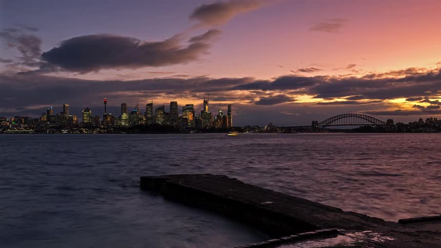 Timelapse of boats moving across a harbor with a city skyline and bridge in the background