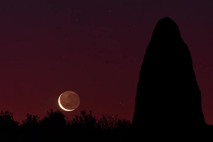 Moon sets as the trees and a tall rock formation stand against a darkening red tinted sky