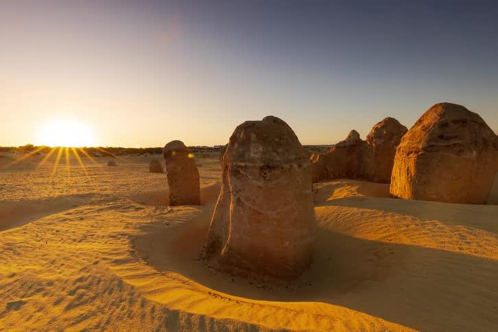 A wide time lapse shot captures the sun setting behind weathered limestone formations in a vast desert while long shadows stretch across the rippled yellow sand under a clear sky that transitions from bright gold to deep blue