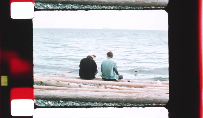 Two people sit side by side on a beach looking out at the ocean framed within a vintage film strip