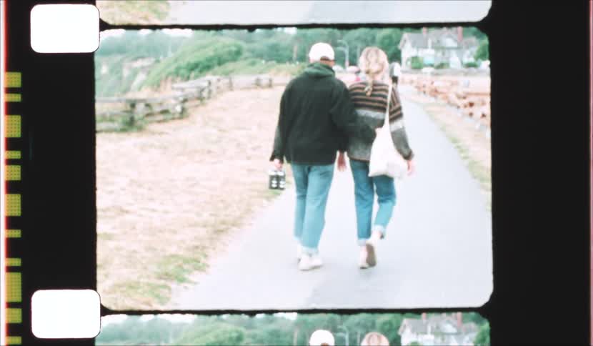 A man and woman walk away from the camera along a paved coastal path next to a wooden fence