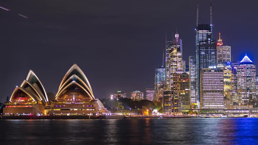 A time lapse shows the sydney opera house and the city skyline at night with boat traffic in the harbor