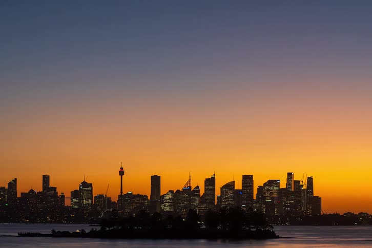 A dark silhouetted city skyline rises behind a calm body of water and a small treecovered island while a vivid orangetopurple sunset glows across the sky highlighting a tall observation tower and modern highrise buildings