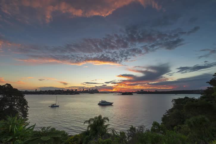 Sunset over bay with solitary sailboat and distant city skyline beneath scattered clouds
