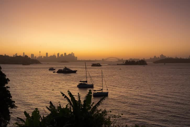 A series of boats sailing along the coastline with a cityscape in the background as dusk turns into night