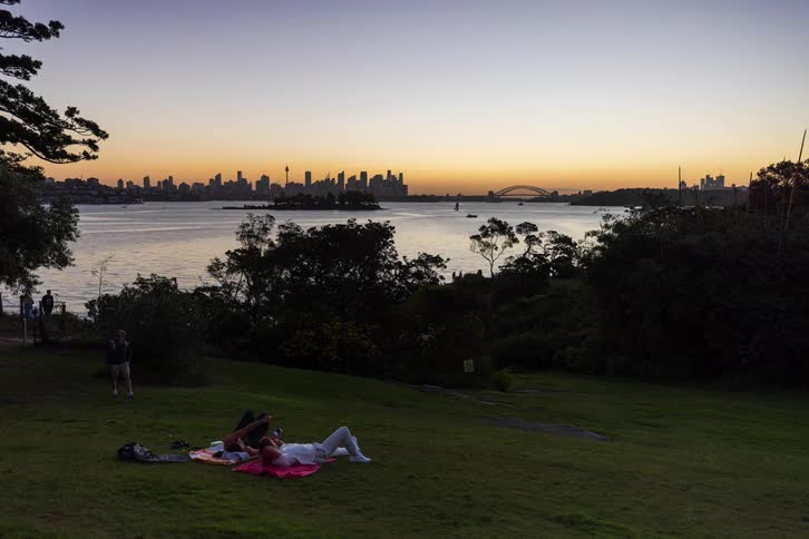 Two people lying on blanket on grass watch sunset facing bay and city skyline