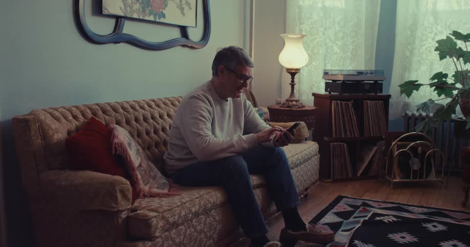 Man sitting on sofa uses phone in living room with lit lamp and record shelf nearby