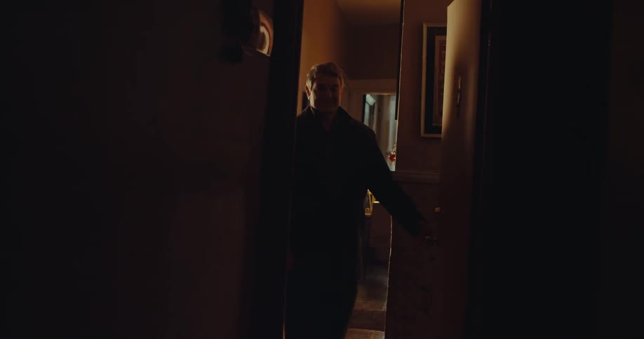A man with grey hair wearing a dark jacket and collared shirt walks through a doorway from a brightly lit room into a dark hallway of a house