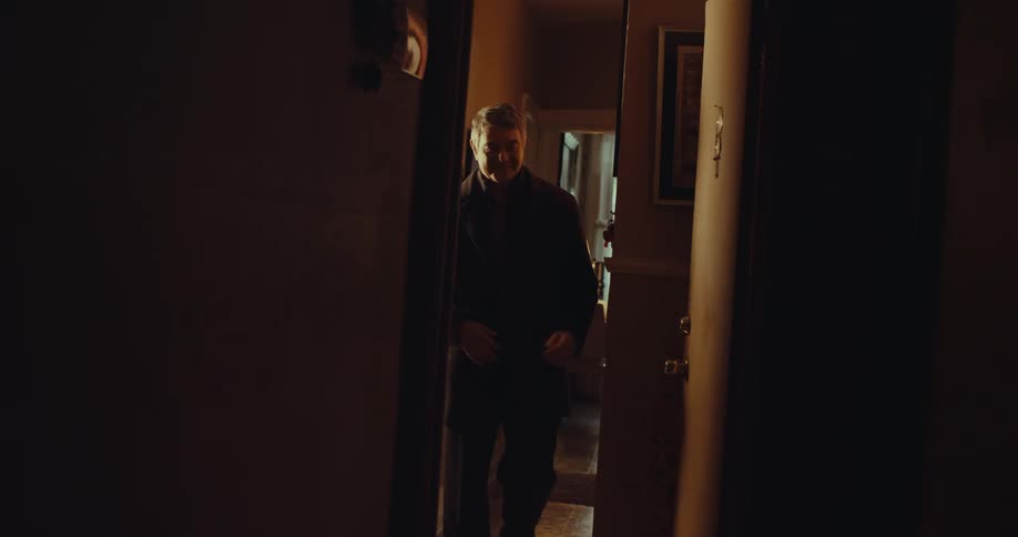 A man stands in the doorway of a warmly lit home hallway adjusting the front of his dark coat as he prepares to leave with framed artwork and soft amber lighting