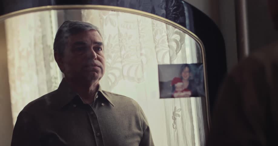 A man with grey hair stands before a mirror carefully fastening the buttons on his buttondown shirtthe reflection shows a photograph of a woman and child tucked into the frame of the mirror beside him