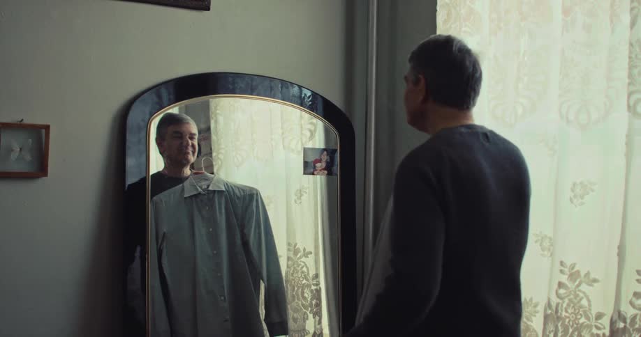 A man stands before a mirror holding up two different buttondown shirts on hangers to compare them he carefully evaluates his reflection appearing to decide which garment to wear for the day