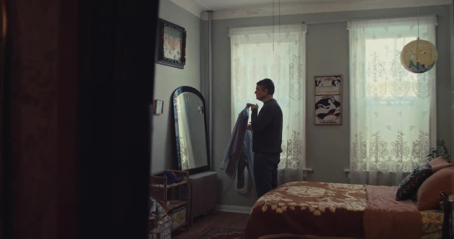 A man stands in a bedroom holding a blue shirt while looking into a large mirror he adjusts the garment and checks his reflection before starting to put it on